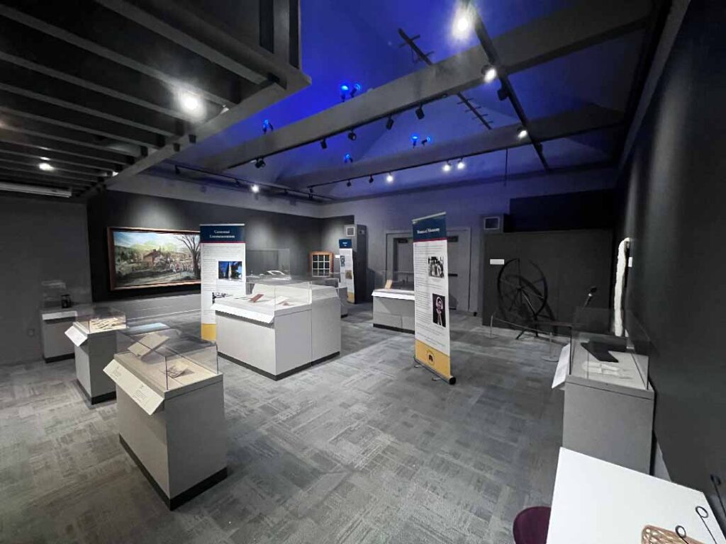 Museum gallery with grey carpeting and white display tables covered with glass tops over different artifacts. The ceiling light is white with blue highlights
