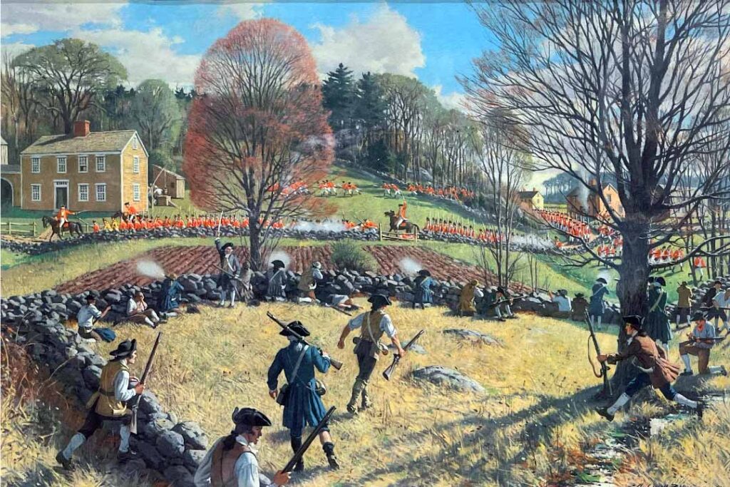In the foreground is a group of colonial militia men dressed in in a variety of clothes - either blue coats, brown coats, or light brown waist coats. The men are hidden behind a wall made of large stones and several are firing muzzle loading firearms. Just beyond the stone wall is a column of British soldiers marching in a perfect line led by officers all wearing bright red coats. Beyond the British is a small house and woods.