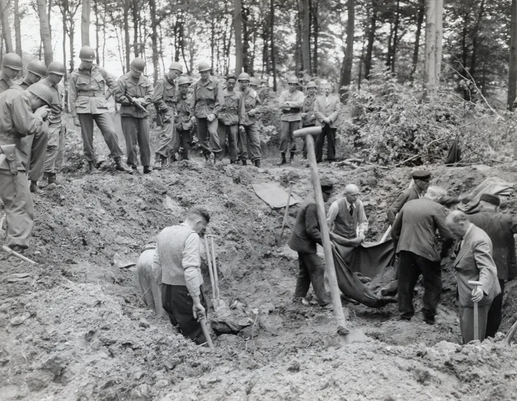 Soldiers of the 194th Glider Infantry Regiment guard Nazi Party members dressed in their Sunday best, forcing them to exhume the bodies of thirty-five people dumped in a bomb crater near Duisberg, Germany, 31 May 1945. (National Archives) 