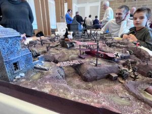 Image is of a miniature game board that illustrates a World War I trench. It includes small Soldier figures, barbed wire, trees, buildings, and planes.